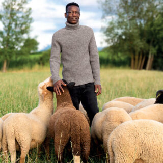 Homme portant un pull col roulé en laine dans une prairie entouré de moutons.